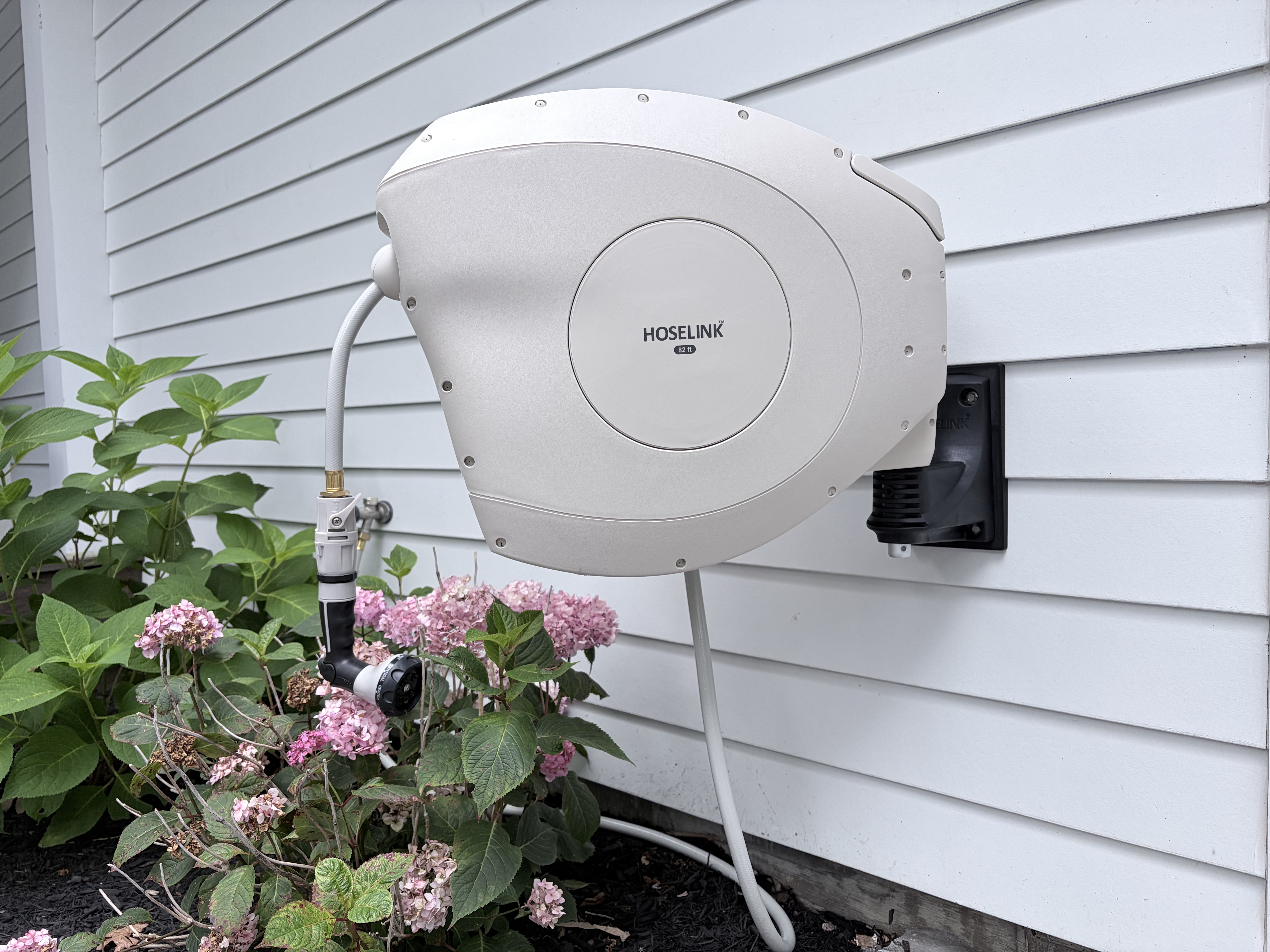 Hoselink retractable hose reel mounted flush on white lap siding above blooming hydrangeas